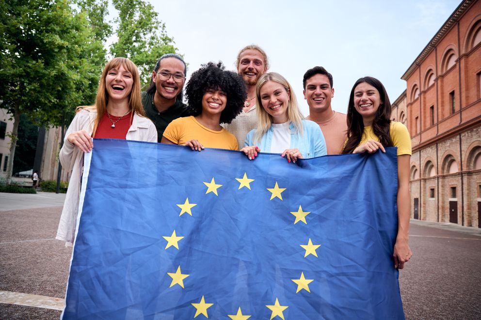 Gruppe junger Lehrkräfte hält Europa-Flagge in Altstadtkulisse hoch